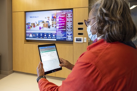 Poppy Bass, senior project manager in Penn Medicine IS, looks at an iPad showing patient chart information available in the Pavilion, while standing in a patient room with a large TV screen in the background.